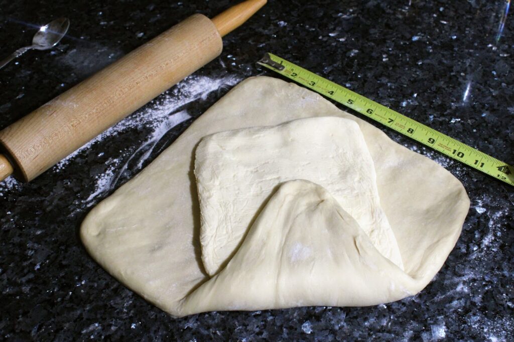 A 8x8-inch square of butter inside of a 12x12-inch square of dough. There is a ruler and a rolling pin in the photo.