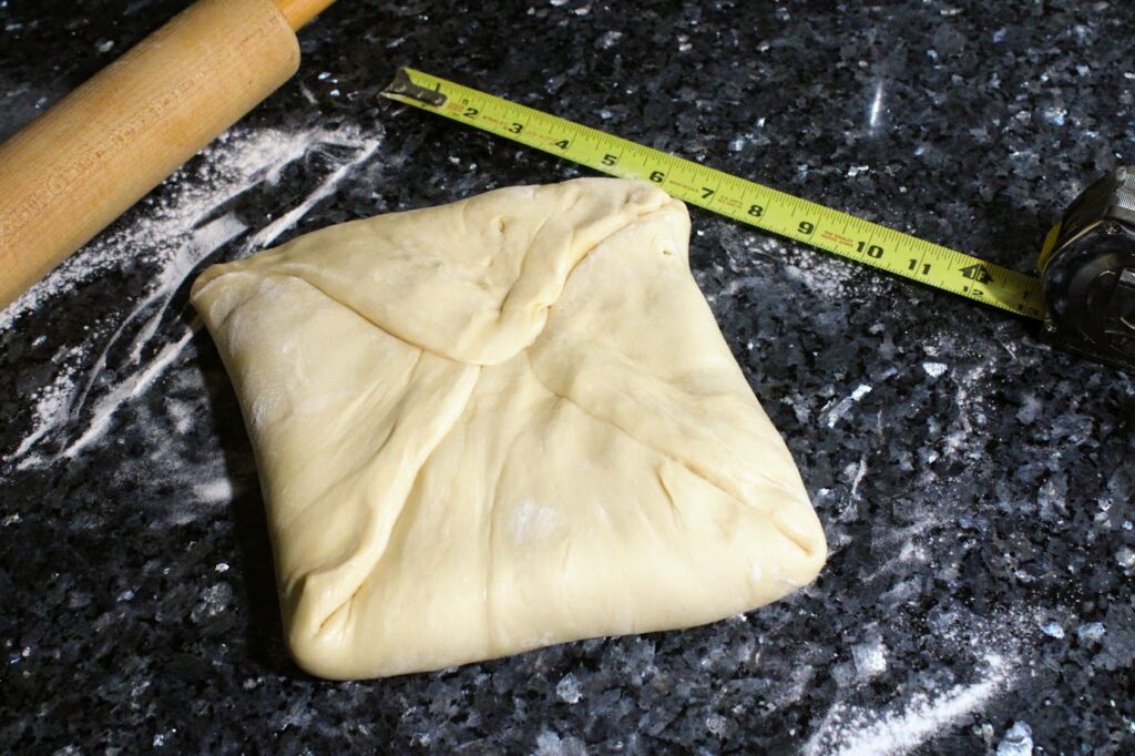 Dough wrapped like a letter around a square of butter. A rolling pin and tape measure are visible.